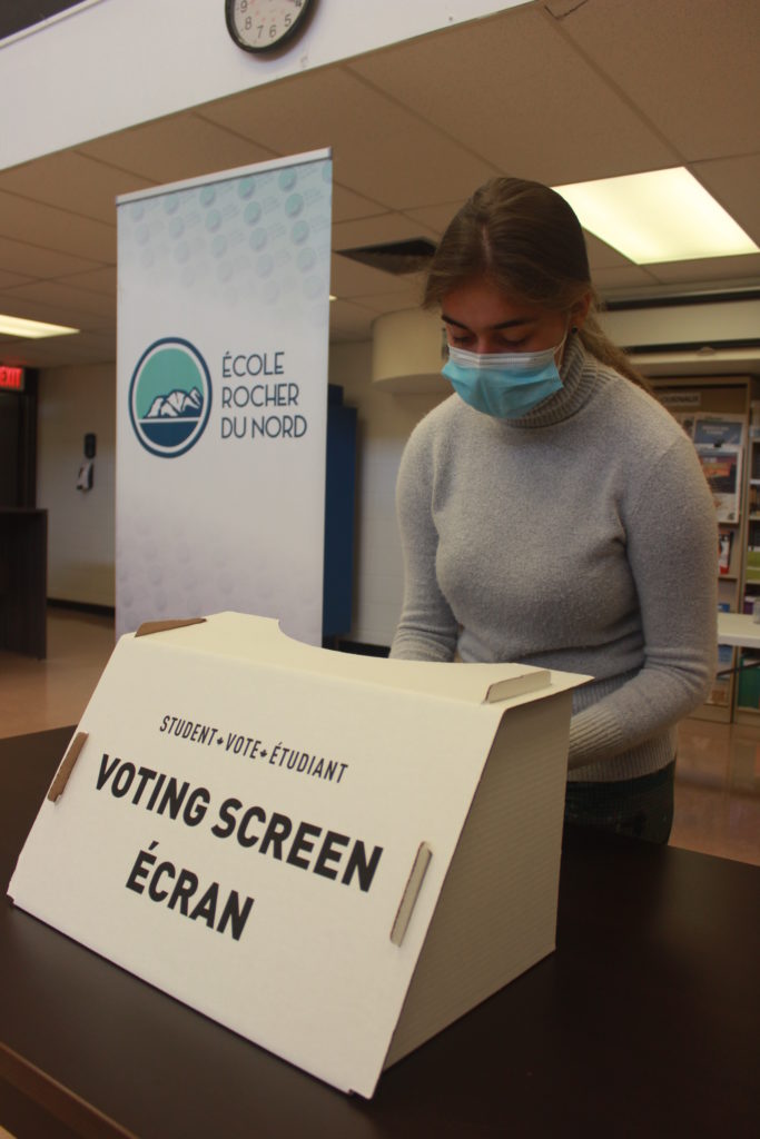 Une étudiante est à une urne ou est écrit «student vote étudian» et «voting screen écran» pour participer au vote étudiant. A sa droite est une bannière qui lit le nom de son école «École Rocher du Nord»