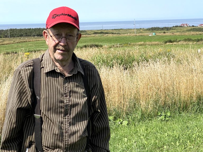 Portrait de Howard Farrell, il est à l'extérieur: derrière lui est un grand espace ouvert de gazon et herbe, plus loin l’océan.