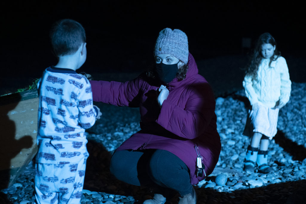 À gauche est un petit garçon qui est de dos a la caméra, au centre une femme avec un gilet violet qui lui parlent, a droite une petite fille. C’est la nuit et les trois sont illuminés de lumière bleue. 