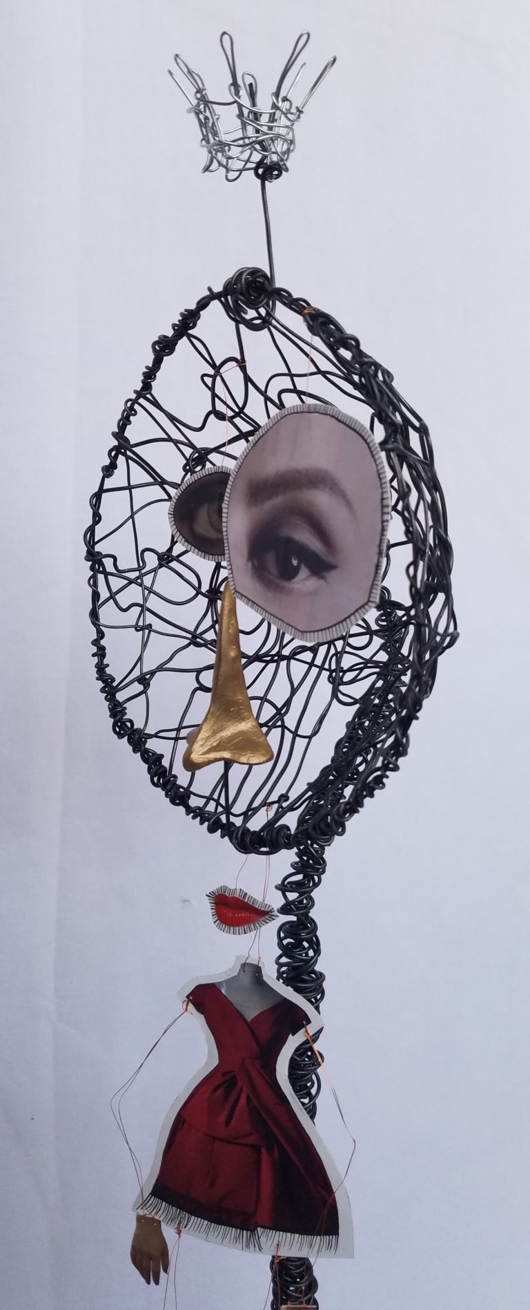 Sculpture de Eveline Ross-Phaneuf. Fait de métal tordu avec morceaux de papier (avec deux yeux, un nez, une bouche, une robe rouge, deux mains, et deux souliers) suspendu devant.