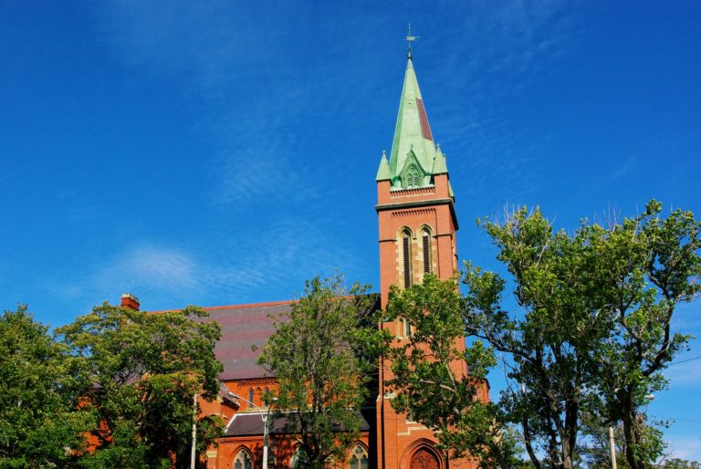 Une église de briques rouges, avec des arbres pleins de feuilles vertes en avant, avec un ciel bleu au fond.