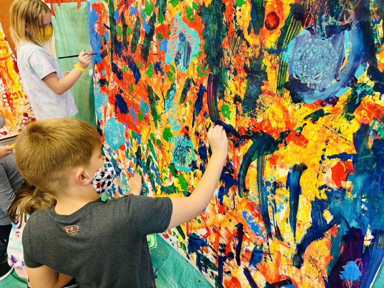 Une peinture abstraite et multicolore fait presque toute l'image. À la gauche est un enfant de dos à la caméra qui contribue au tableau.