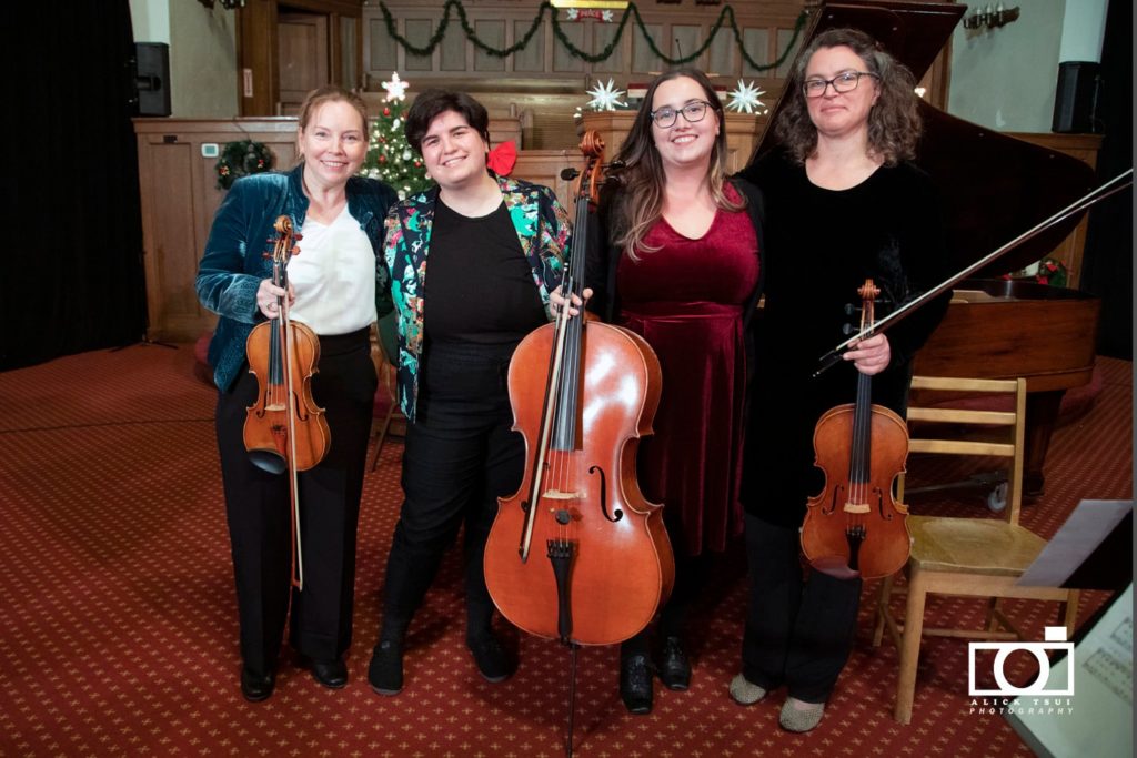 Dans le premier plan, quatre femmes ensemble avec leurs instruments à cord. Dans l'arrière plan, une salle avec des décorations de Noël
