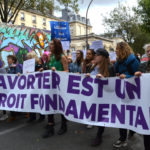 Un groupe de femmes d'âges différents marche le long d'une route en tenant une bannière blanche avec une inscription violette qui dit «avorter est un droit fondamental».