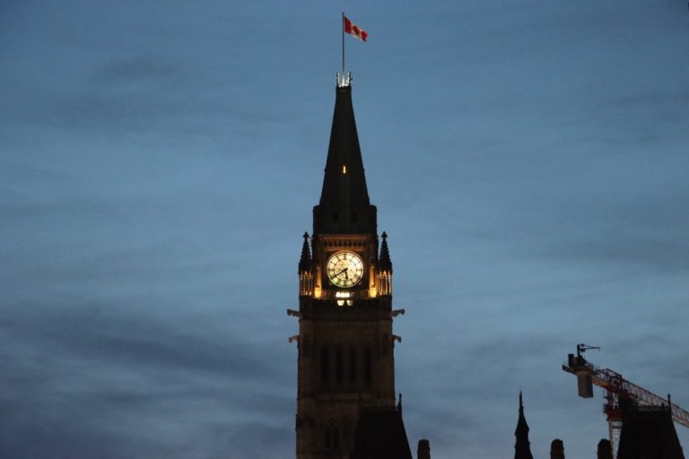 Au centre de l'image se trouve une tour d'horloge. La photo a été prise au crépuscule et, par conséquent, l'obscurité masque les couleurs et les détails de la tour. Une horloge blanche est éclairée au centre. Un drapeau canadien rouge et blanc est sur un mât fixé au point le plus haut de la tour. En bas à droite se trouvent quatre pointes d'autres tours dans l'ombre, et dans le coin se trouve le sommet d'un bâtiment rectangulaire surmonté d'une grue. Le ciel est bleu avec des nuages gris.