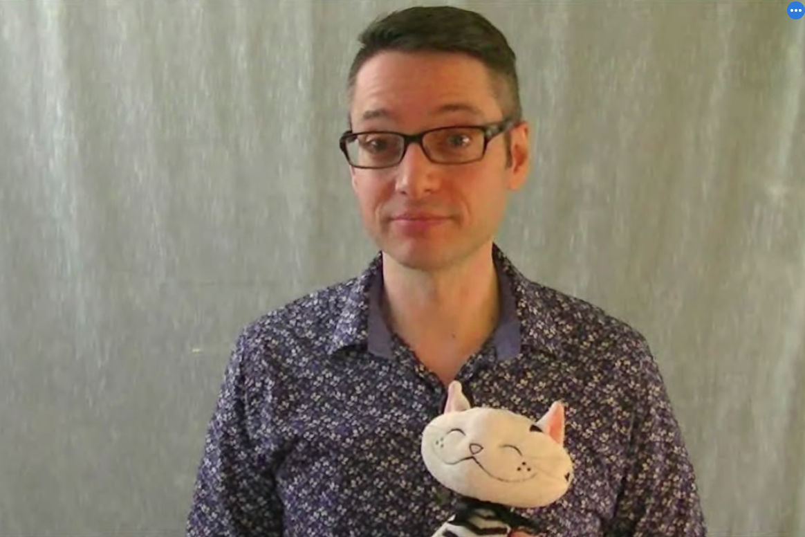Un homme aux cheveux noirs courts fait face à la caméra et tient un chat en peluche noir et blanc. Le chat sourit, les yeux fermés. L'homme porte une chemise bleue et blanche et des lunettes rectangulaires à monture noire épaisse. Il y a une toile de fond grise en arrière-plan. 