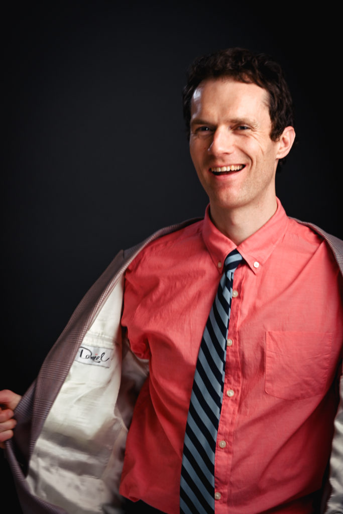 Un portait du haut du corps d’un jeune homme qui porte un veston gris, un chandail rose et une cravate raillée. Il sourit vers l’objectif. 