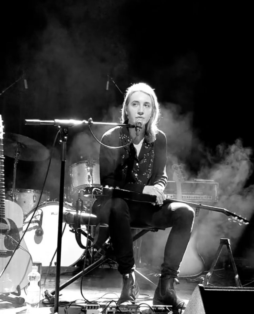Photo en noir et blanc d'une artiste sur scène pendant un concert. Elle est assise sur un petit banc noir avec une guitare sur ses jambes et a un microphone sur sa gauche.