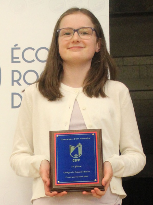 Une jeune fille est debout devant une affiche blanche et tient une plaque bleu foncé. 