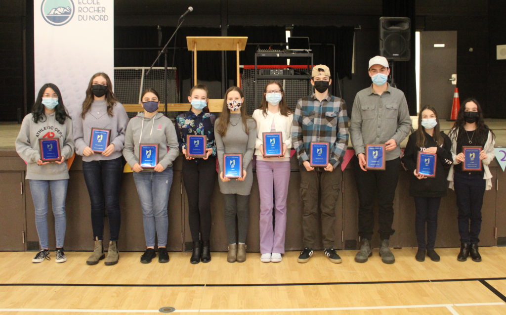 Les jeunes lauréat.e.s du concours d'art oratoire se présentent devant une scène pour une photo de groupe. Tout le monde tient une plaque bleu foncé avec une bordure en bois et porte un masque.