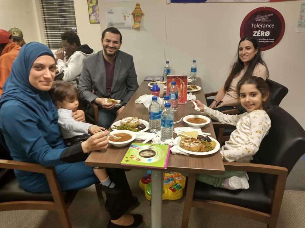 Une famille composée de deux femmes, d'un homme et de deux jeunes enfants est assise à une table en bois brun avec de la nourriture. Le groupe sourit à la caméra.