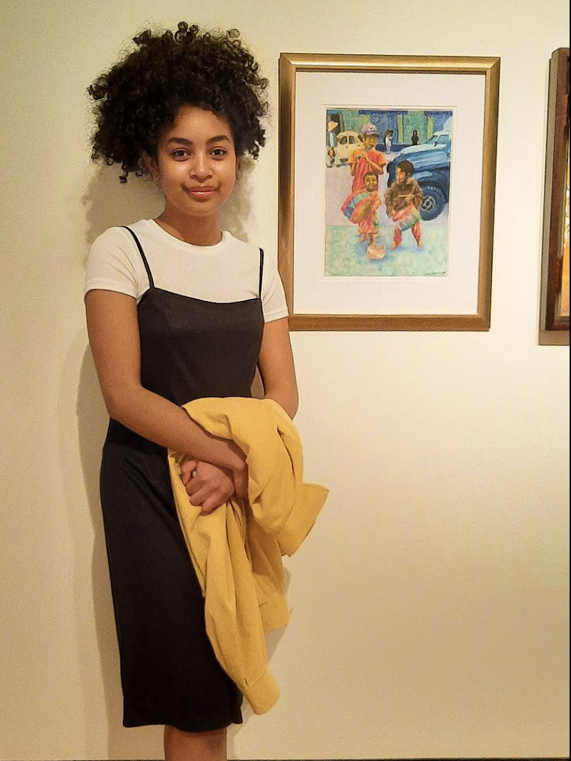 Une jeune fille debout à côté d’un oeuvre d’art accroché sur un mur blanc. Elle porte une robe noire avec un chandail blanc en dessous et teint un vêtement jaune dans ses mains. 