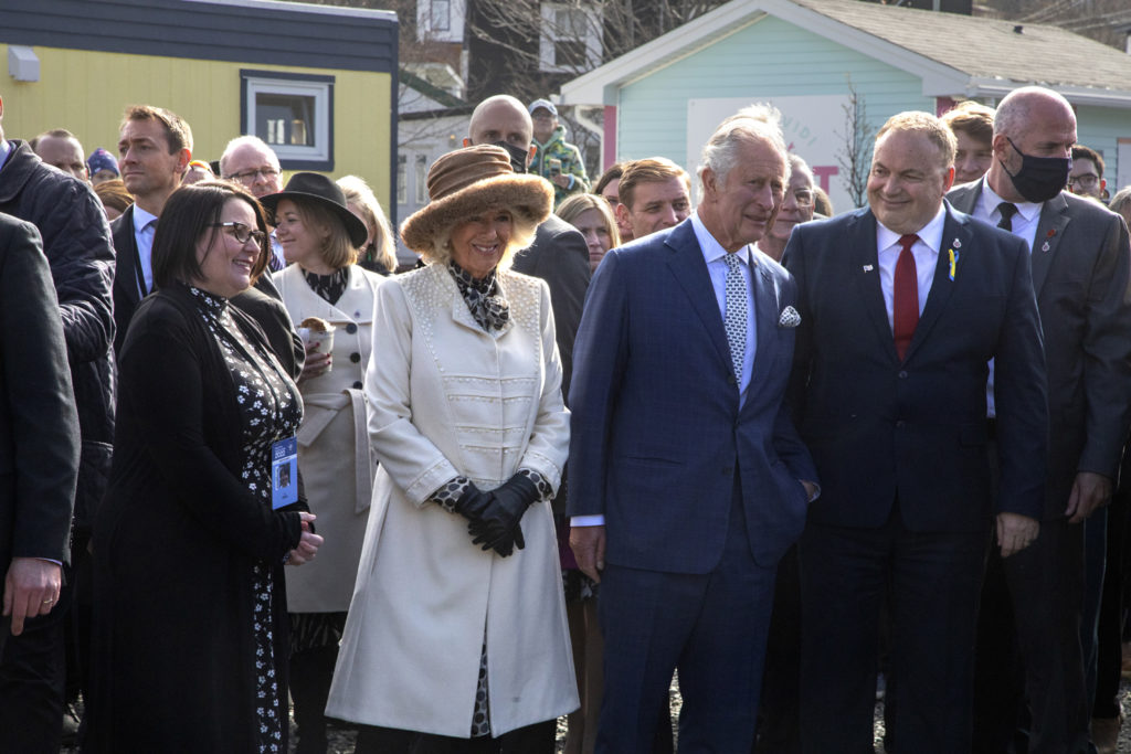 Le prince Charles et la duchesse Camilla d'Angleterre au centre d'un groupe de personnes. Ils regardent vers la droite de la photo. À l'arrière-plan se trouvent deux petits bâtiments, l'un vert et rose sur la droite et l'autre bleu et jaune sur la gauche.