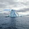 Un petit bateau avec quelques passagers est très proche du côté gauche d’un grand iceberg. Le ciel est nuageux et l’eau de l’océan est bleu-gris.