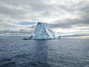 Un petit bateau avec quelques passagers est très proche du côté gauche d’un grand iceberg. Le ciel est nuageux et l’eau de l’océan est bleu-gris.