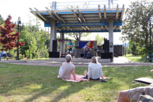 Deux personnes sont assises dehors sur de l’herbe et regardant des musiciens sur une scène.
