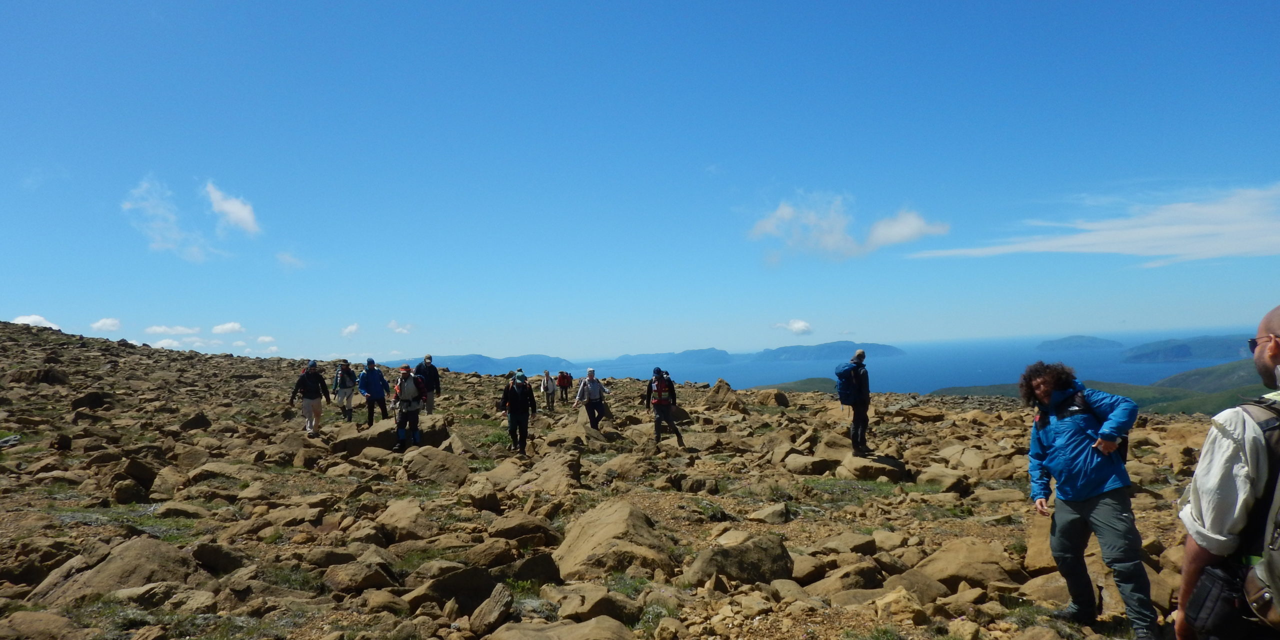 Un groupe de personnes font une randonnée, ils marchent sur des grandes roches brunes sous un ciel bleu pâle.
