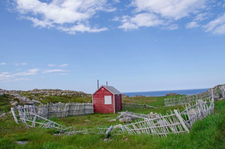 Un petit bâtiment rouge entouré d'une clôture en bois au centre d'un champ de gazon. Le ciel est bleu et l'océan est visible sur la droite.