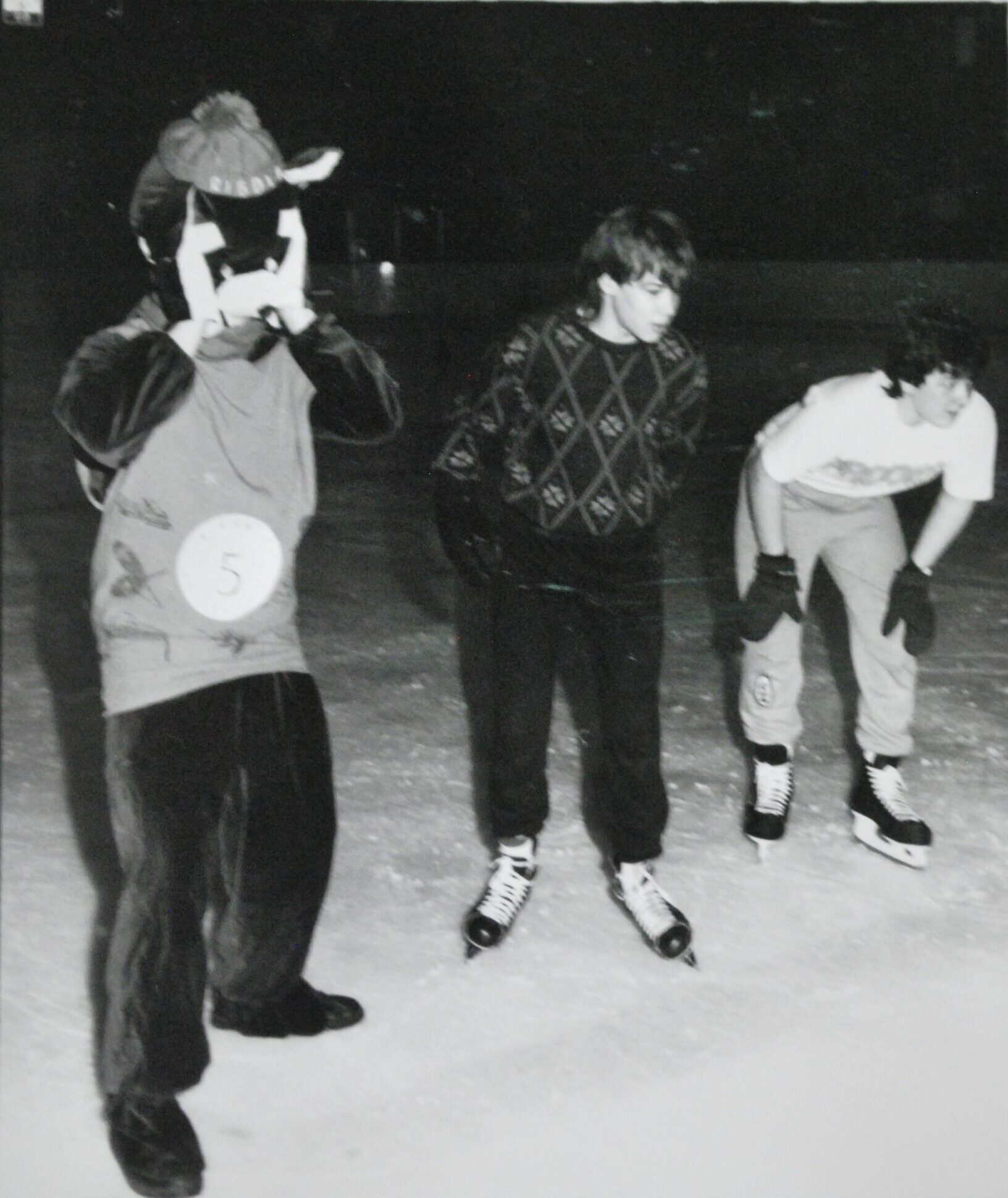 Trois garçons adolescents sur une patinoire le soir. Celui de gauche porte un costume de mascotte.
