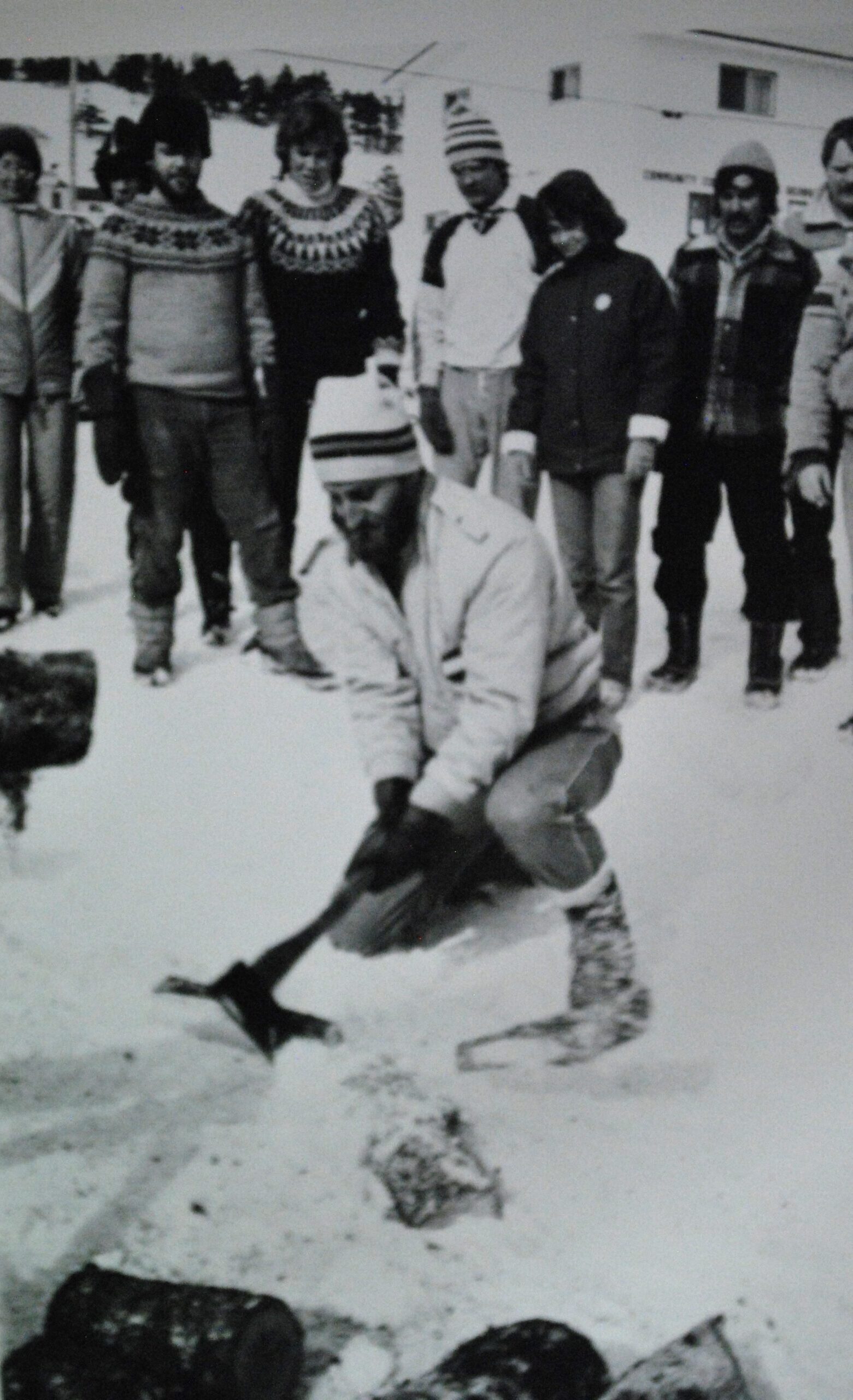 Une photo noire et blanc d'un homme qui frappe un morceau de bois avec une hache en avant d'un group de spectateur. C'est l'hiver et tout le monde porte des vêtements chauds.