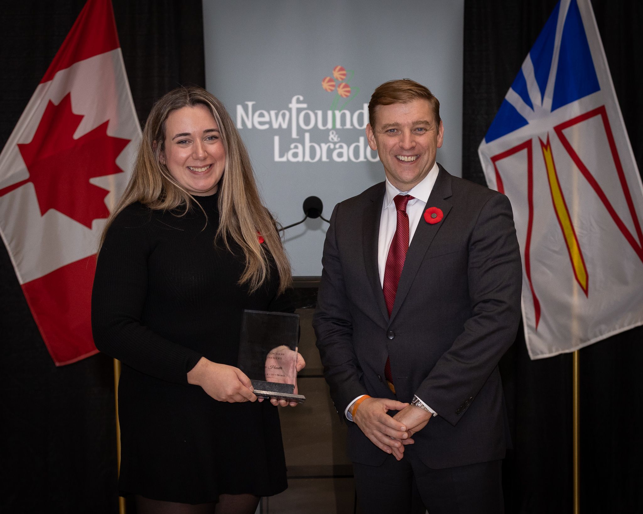 Une femme (à gauche) qui tient un trophée en verre et un homme (à droite) souris vers la caméra. En arrière-plan, de gauche à droite: un drapeau canadien (rouge et blanc), un écran avec «Newfoundland and Labrador» et un drapeau terre-neuvien (blanc, rouge, bleu et jaune).