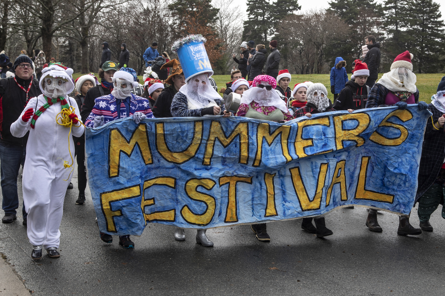 Un groupe de personnes déguiser en mummers marchent dans une rue et tienne une bannière bleue avec les mots jaunes «Mummers festival».