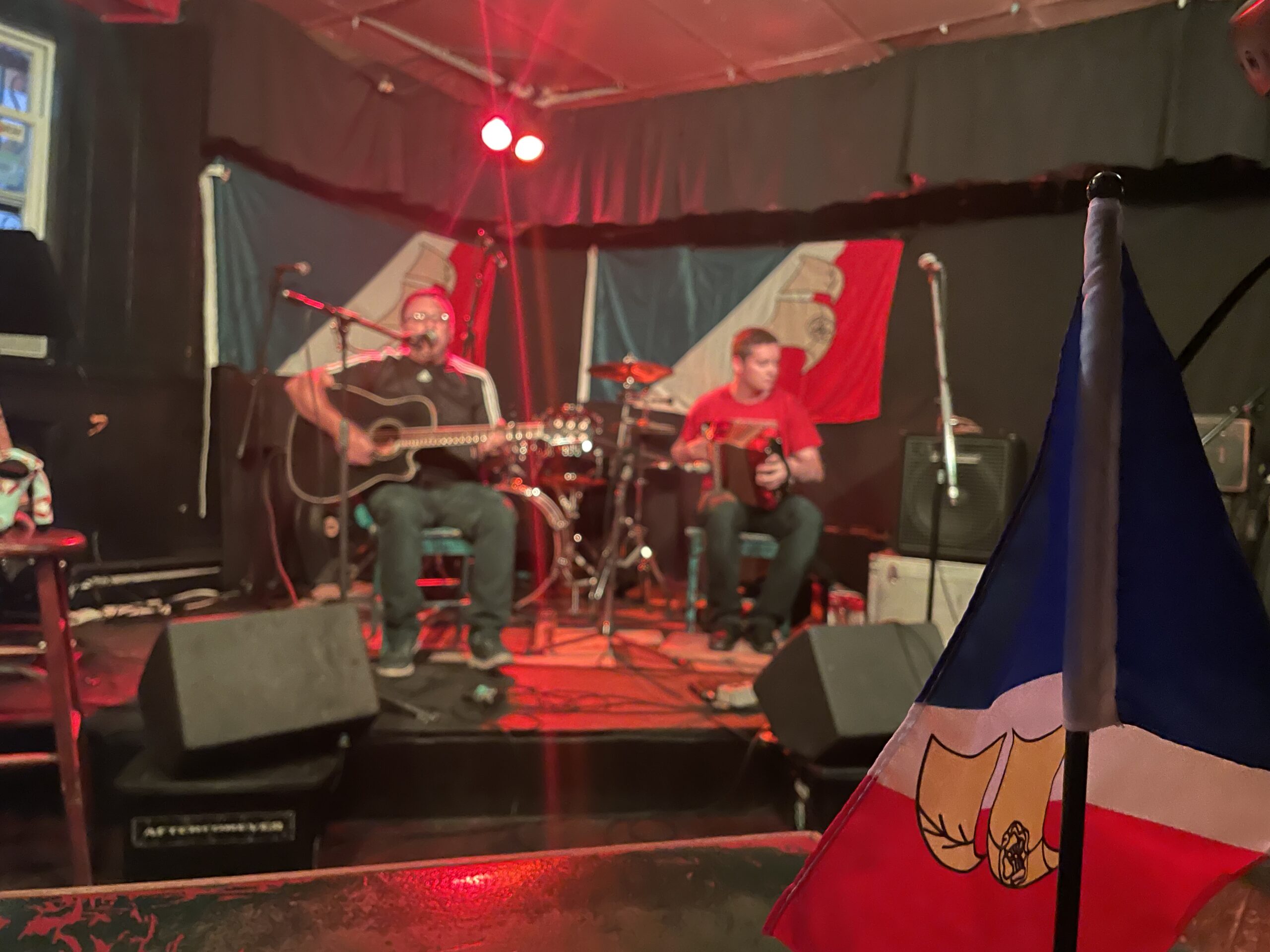 Photo d'un groupe jouant sur scène. Un petit drapeau francophone de Terre-Neuve-et-Labrador se trouve au premier plan à droite.